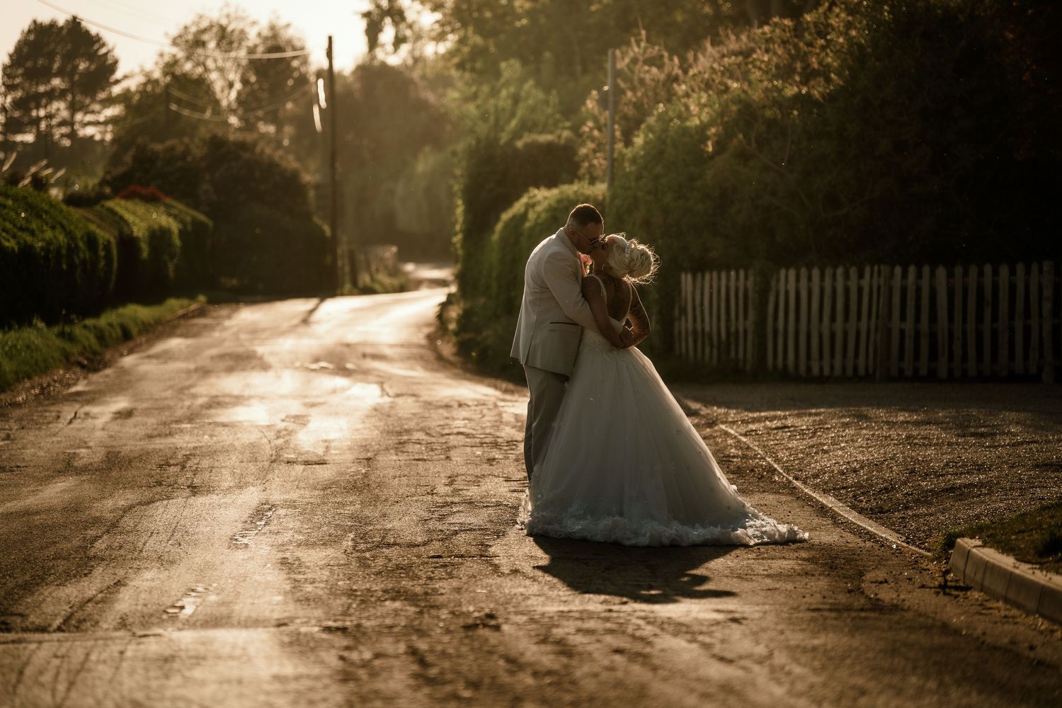 A couple in wedding attire embrace on a sunlit country road lined with hedges at golden hour.