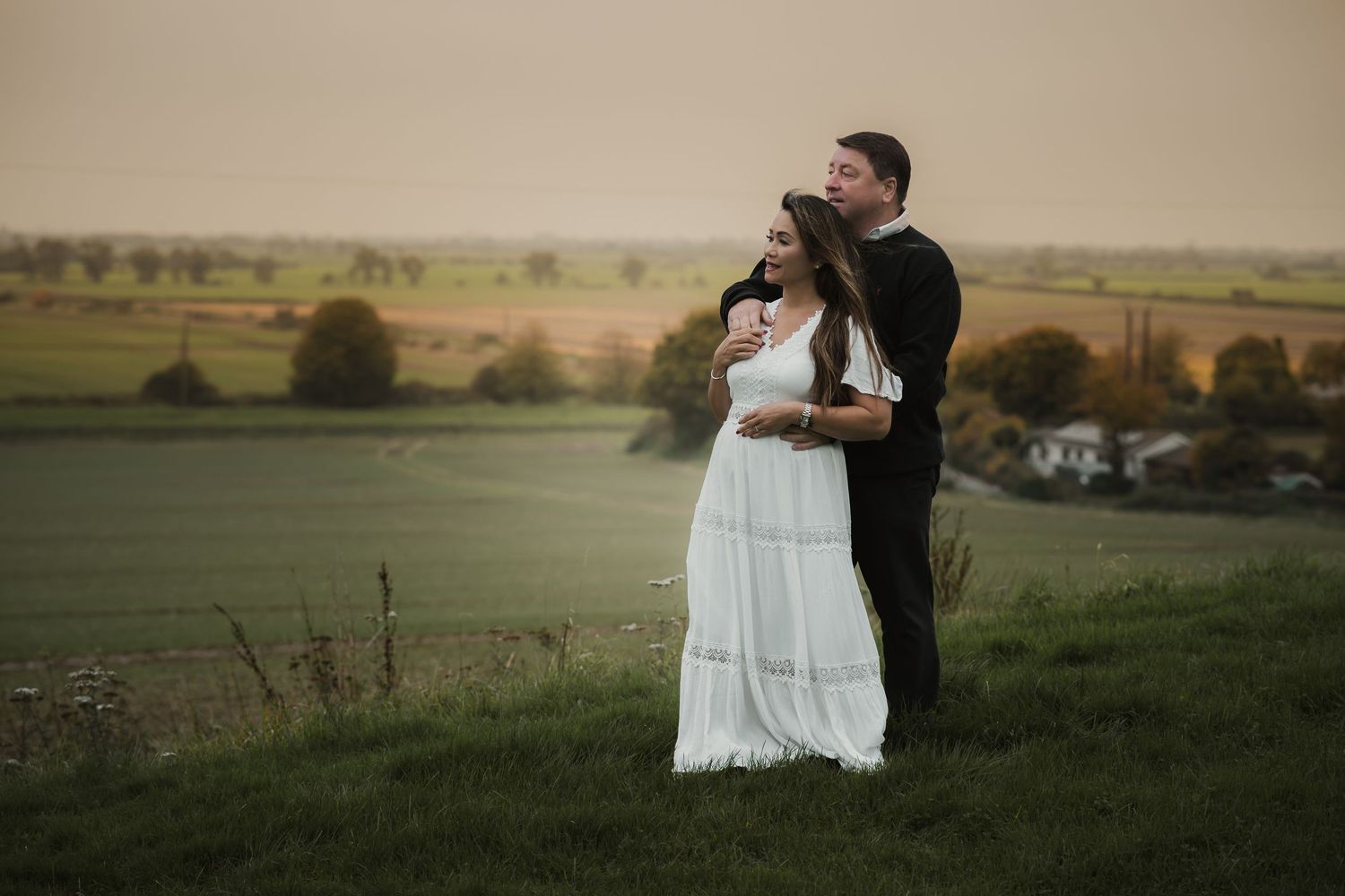 Couple in wedding attire standing on hilltop overlooking scenic countryside.