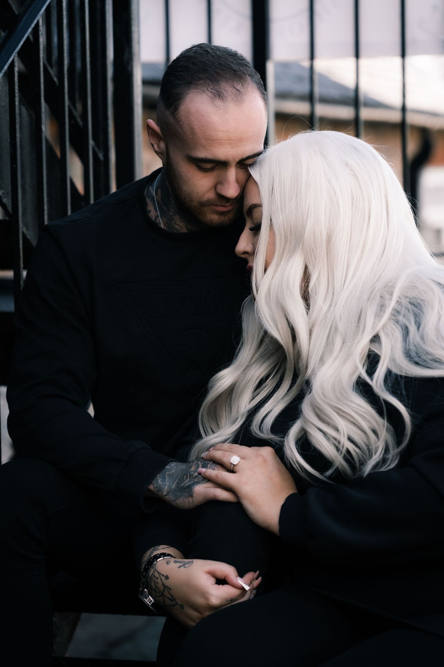 A moody portrait of a couple in dark clothing sharing an intimate moment near metal railings.