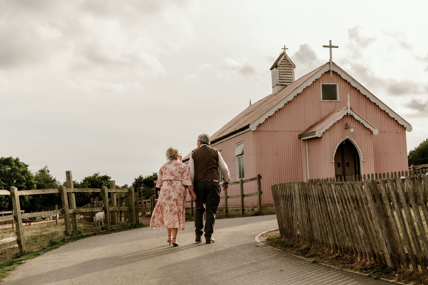 A small pink church building with a wooden cross on top stands against a cloudy sky on a rural hilltop.