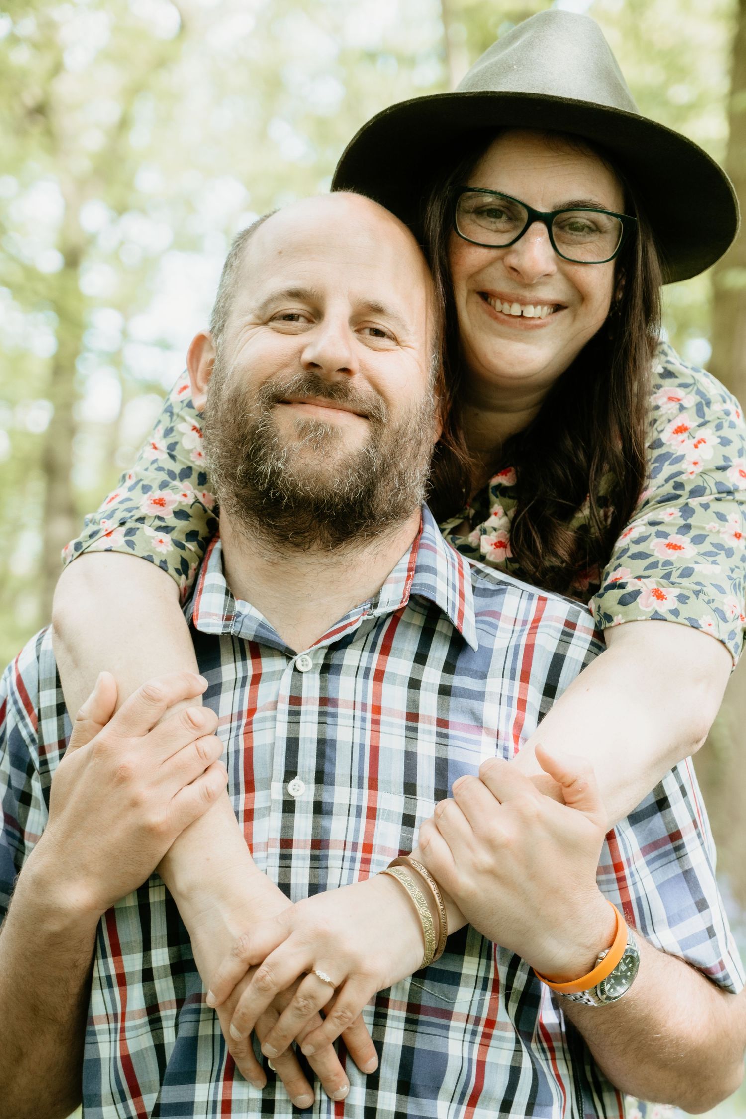 Close-up portrait of a couple embracing outdoors, one wearing a hat and glasses.