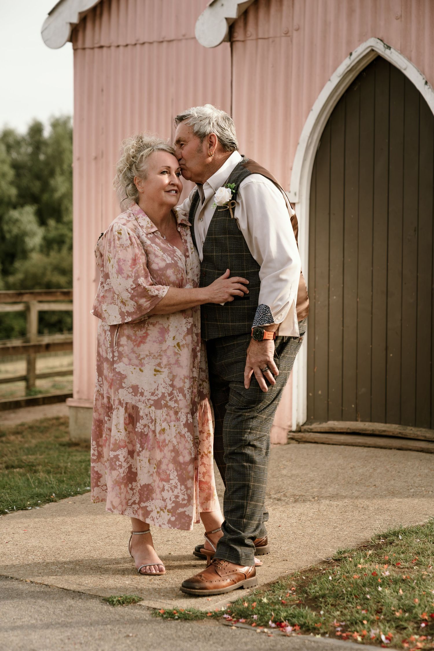 A romantic moment between a couple in vintage-style clothing standing outside a pink barn with an arched doorway.