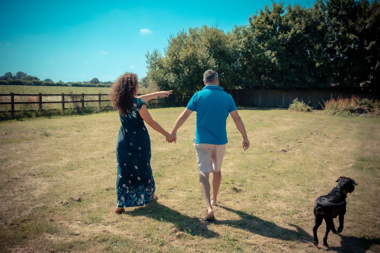 A couple walks hand in hand through a sunny field with their black dog following behind.