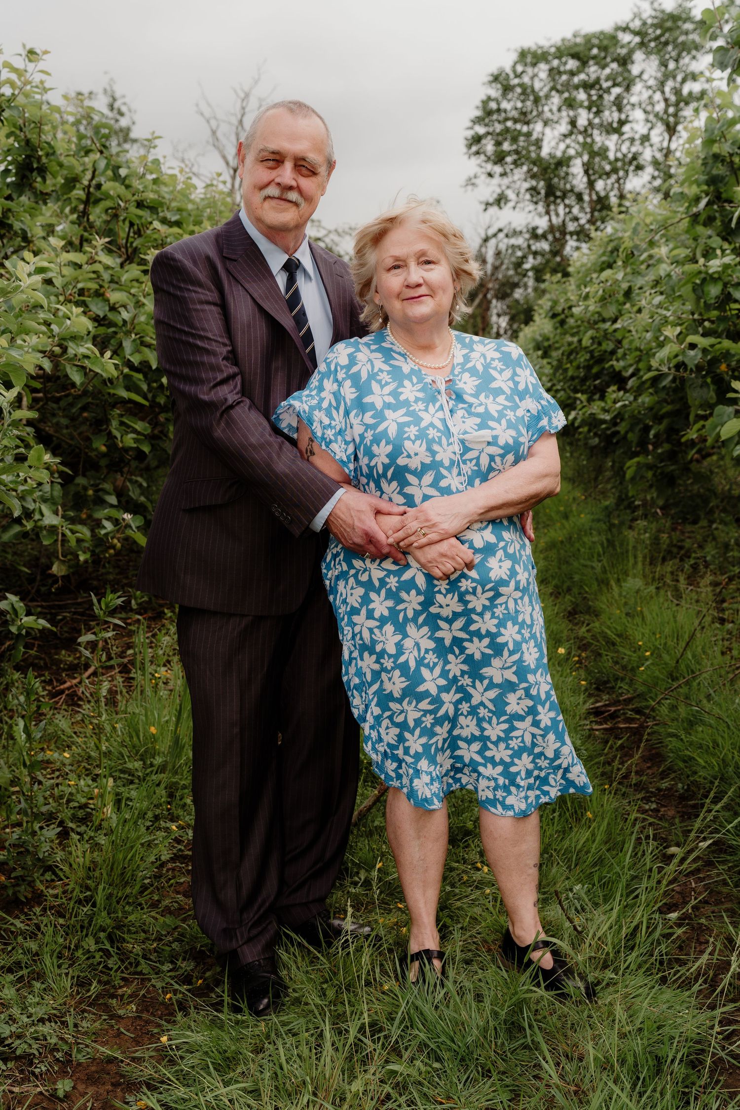 Elderly couple in formal attire pose together in a lush green garden setting.