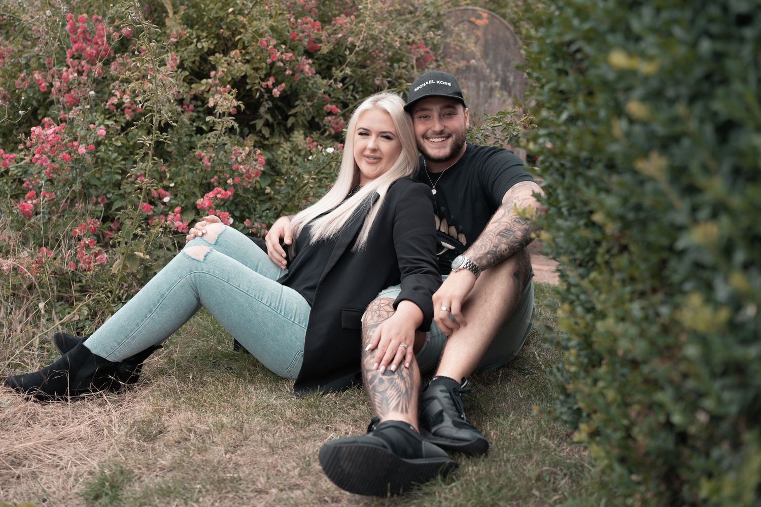 A casual couple relaxing outdoors against flowering bushes in casual attire.