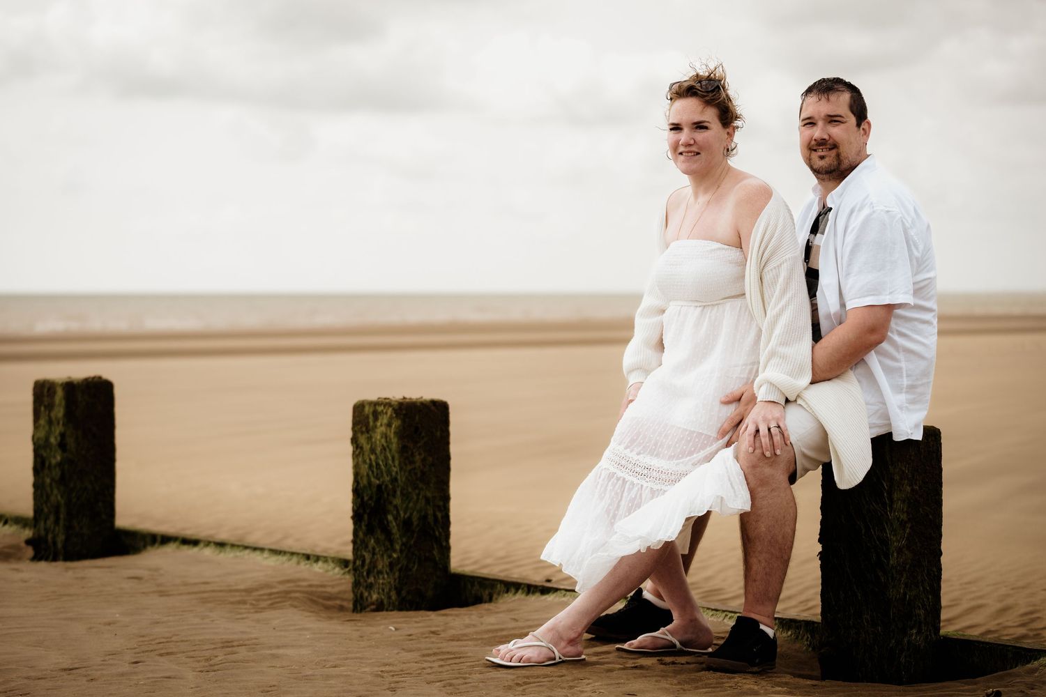 Couple posing by wooden posts on a beach with overcast skies.