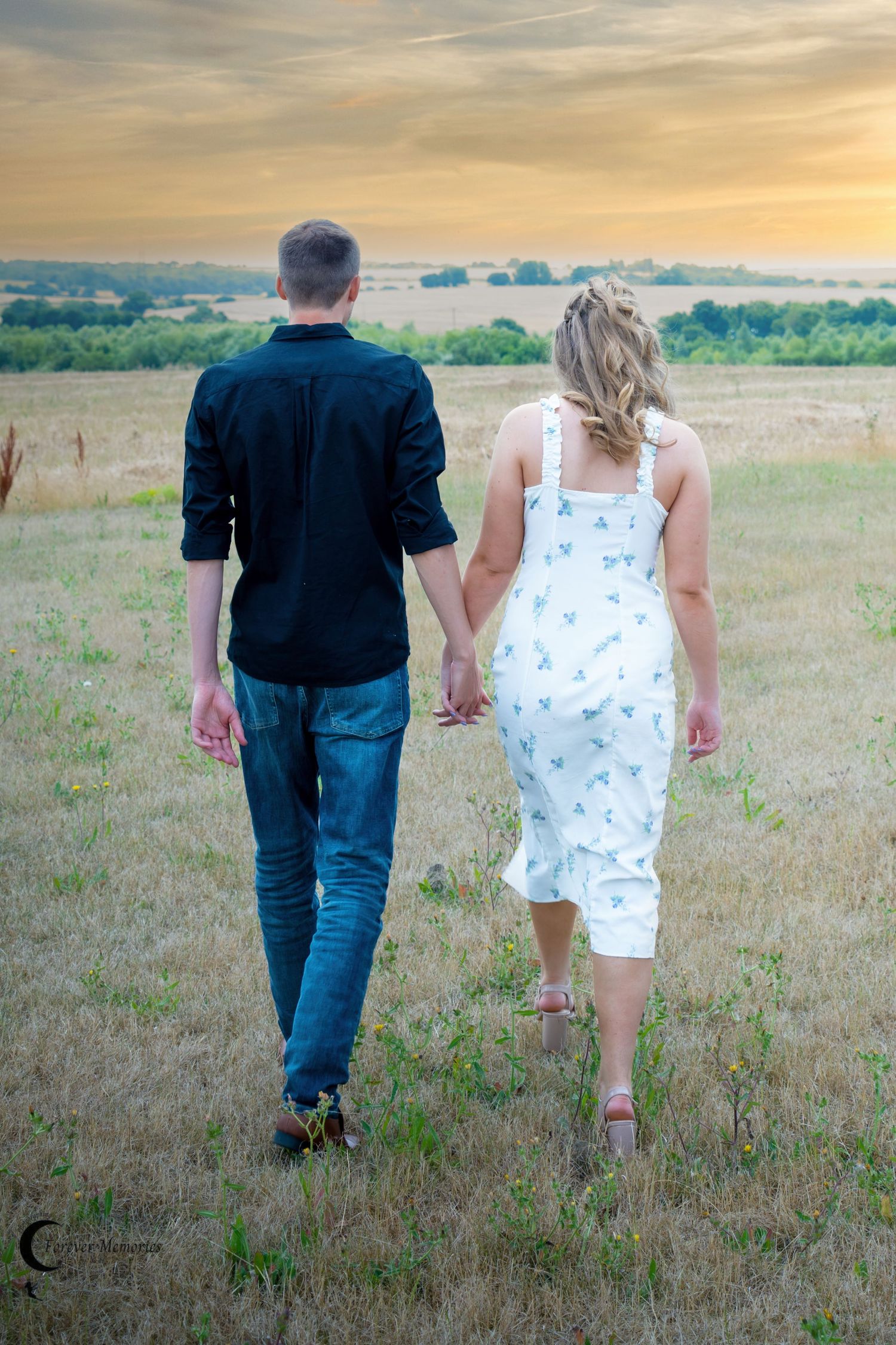 A couple walks hand in hand through a golden field at sunset.