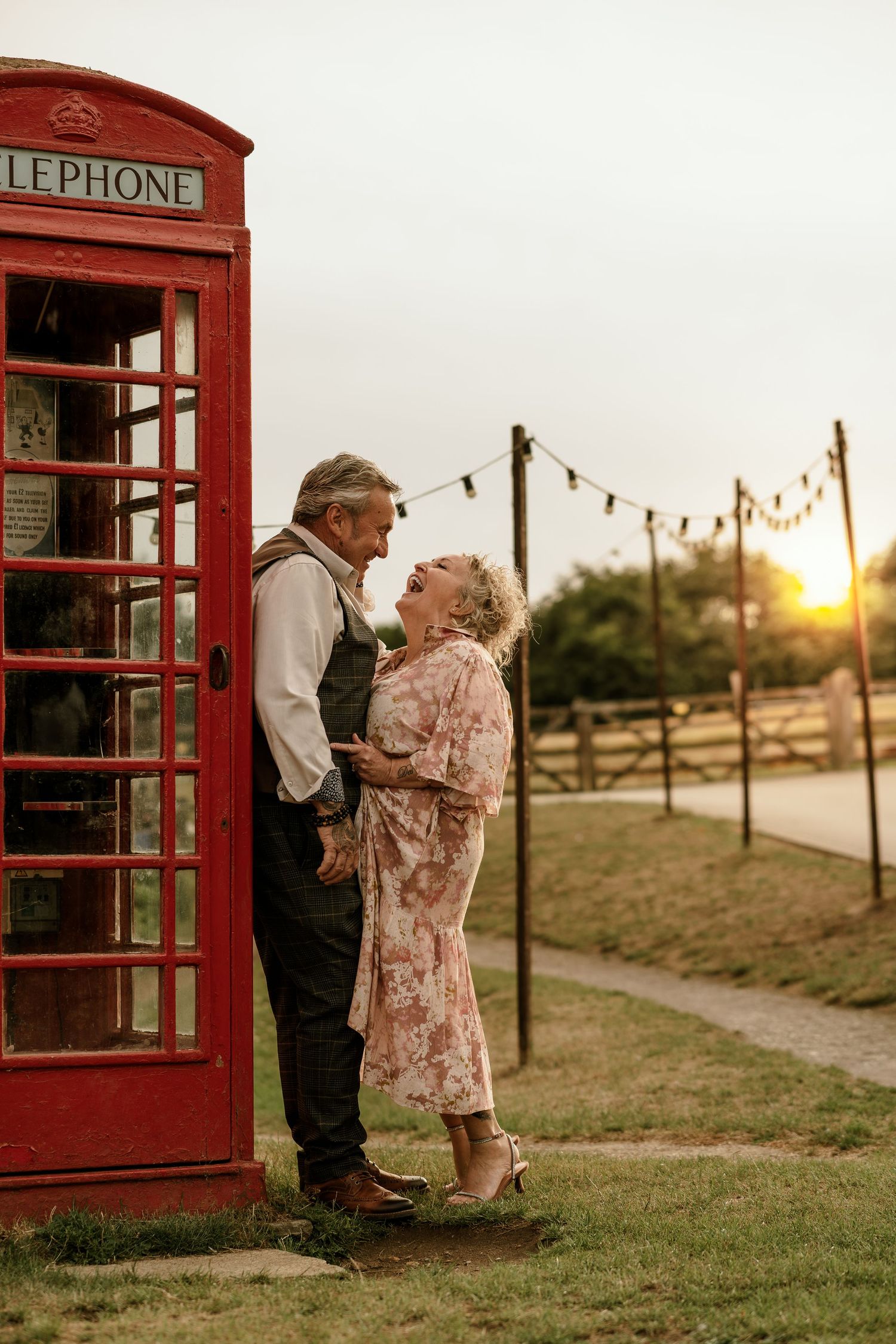 An elderly couple shares a romantic moment by a classic red British telephone booth at sunset with string lights in background.