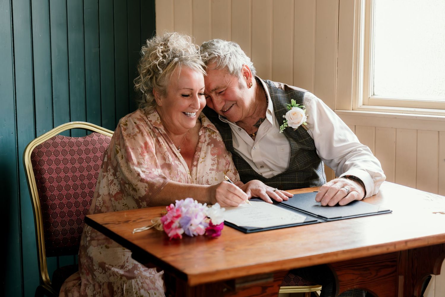 Senior couple reviewing documents together while sitting at wooden table near window.