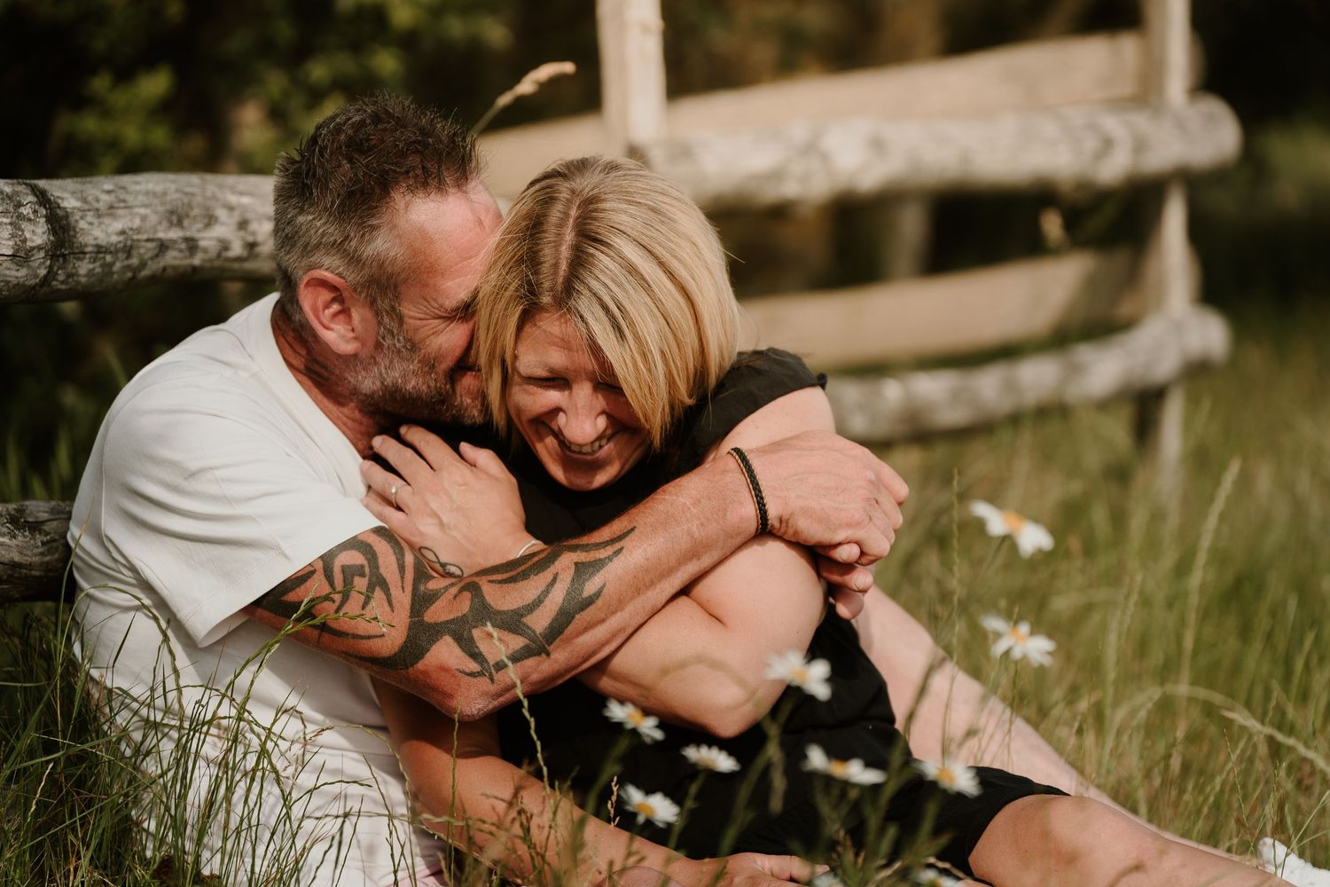 Couple sharing an intimate embrace while sitting by a rustic wooden fence in a meadow.