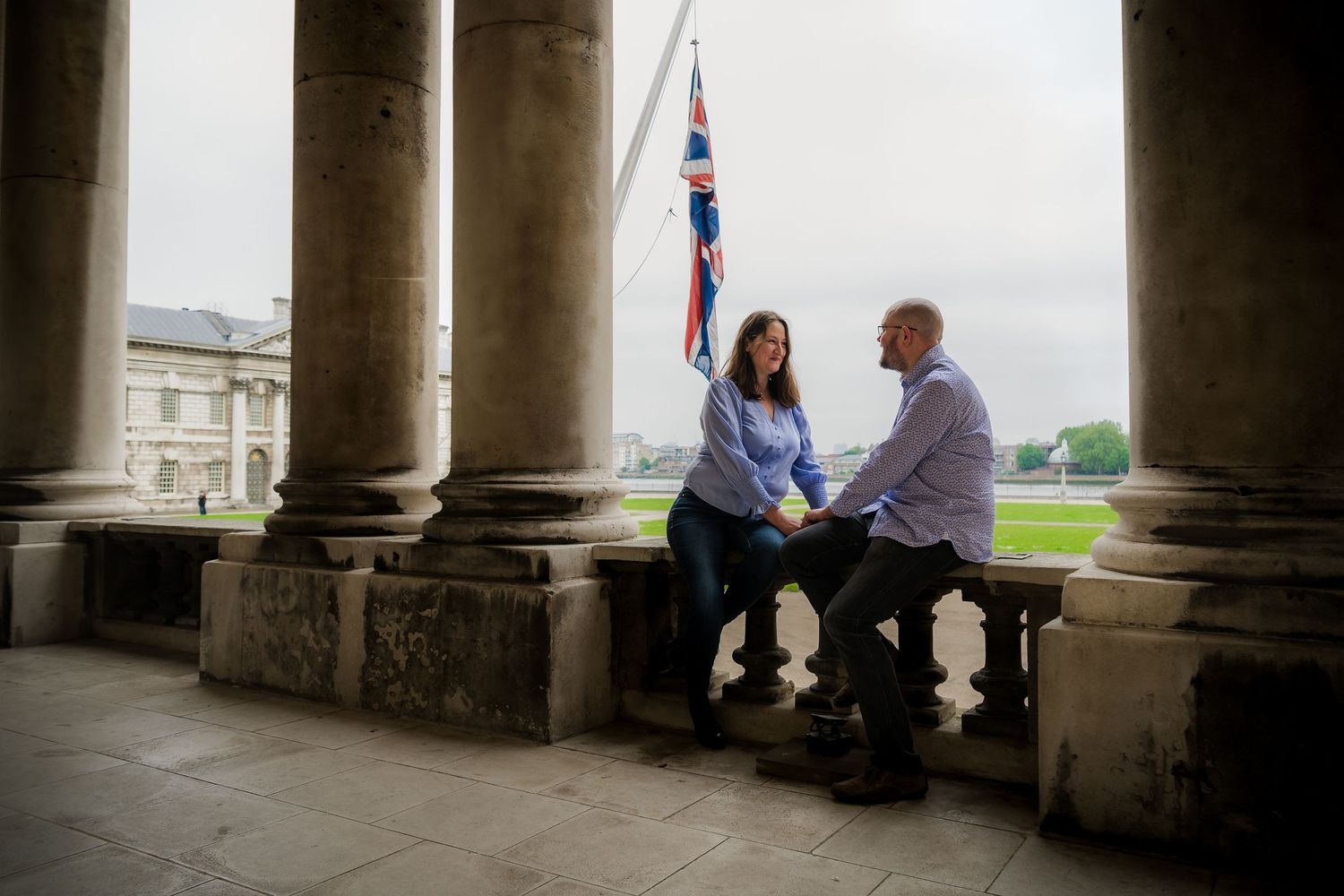 A couple sits between classical columns with a British flag visible in the background.