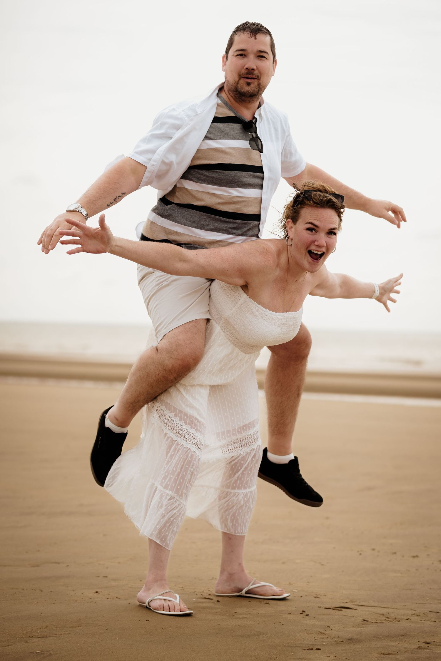 Playful couple in white outfits having fun with piggyback pose on a sandy beach.