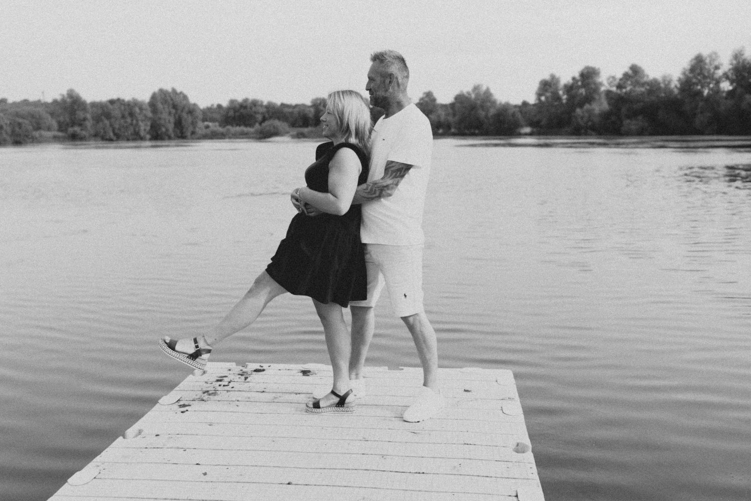 Black and white photo of a couple standing on a wooden dock over a serene lake.