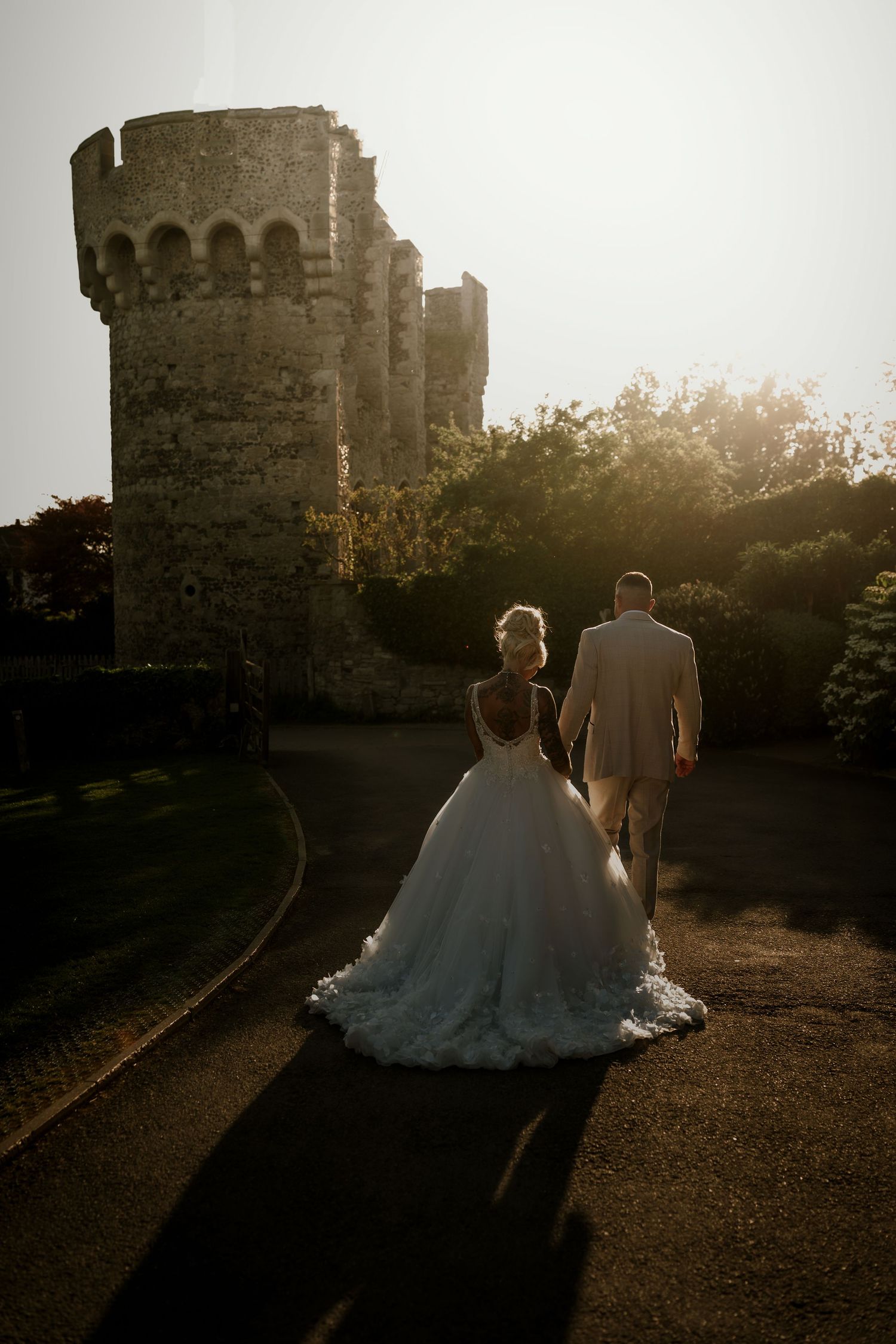 A couple in wedding attire walks along a gravel path at sunset creating a romantic silhouette against castle walls.