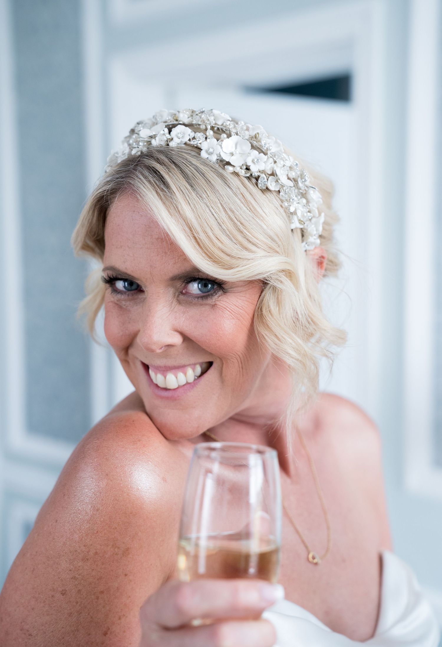 Woman in bridal attire wearing pearl crown and holding champagne glass.