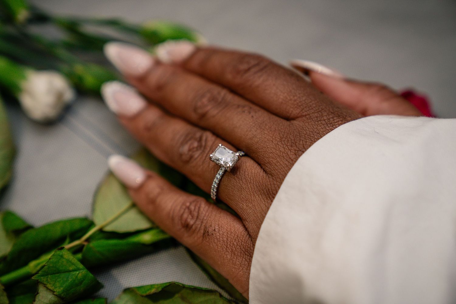 Hand wearing an elegant diamond engagement ring rests against a white sleeve with green leafy roses in the background.