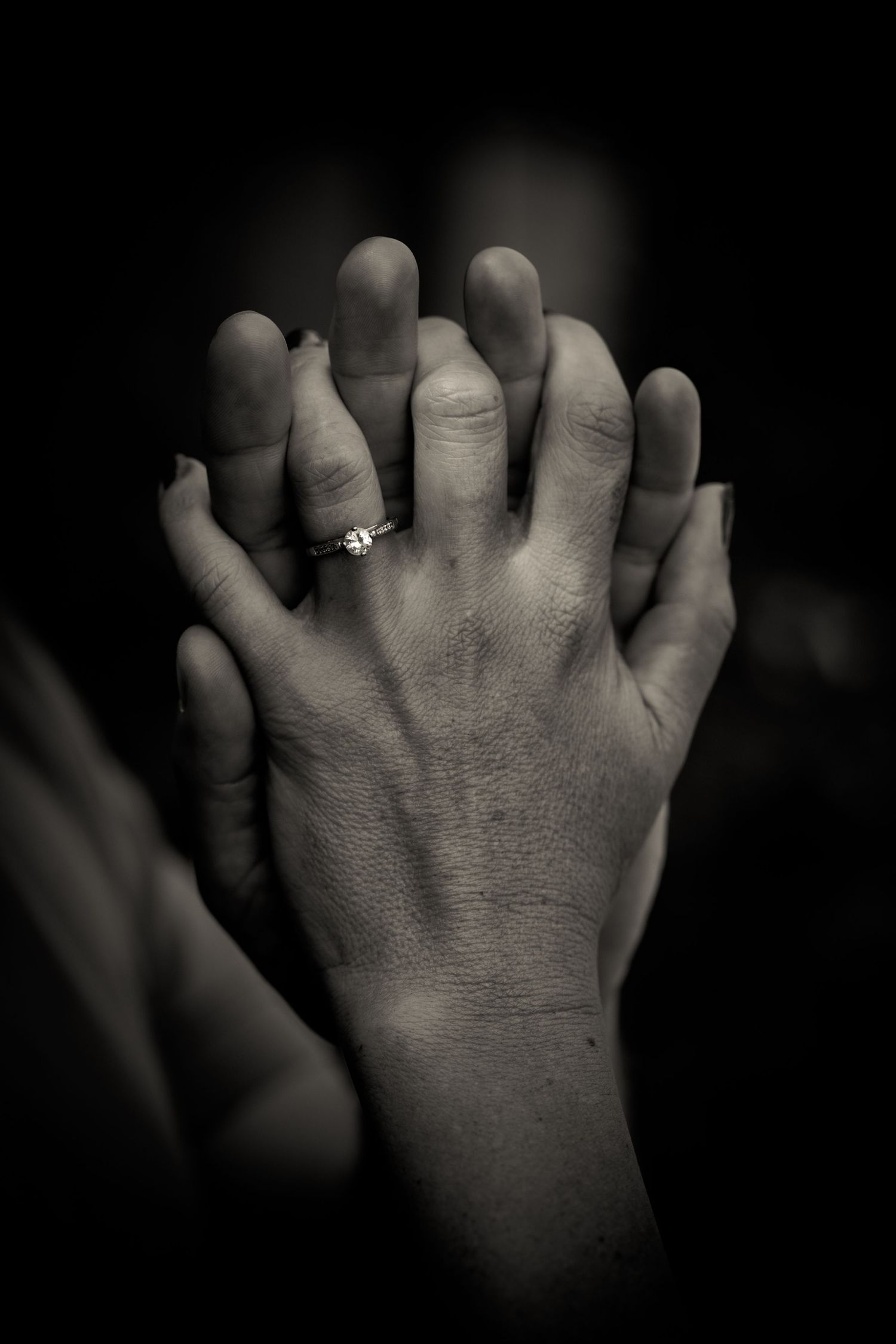 Two hands clasped together in darkness showing an engagement ring.