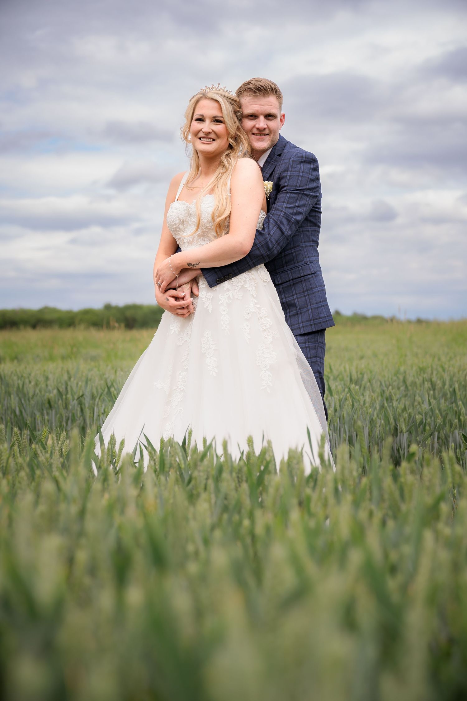 A couple poses together in a lush green wheat field on their wedding day.