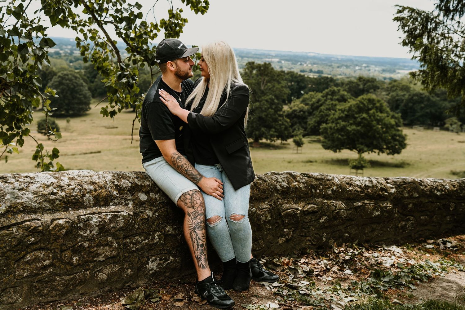 A couple shares an intimate moment while sitting on a stone wall with a scenic countryside view.