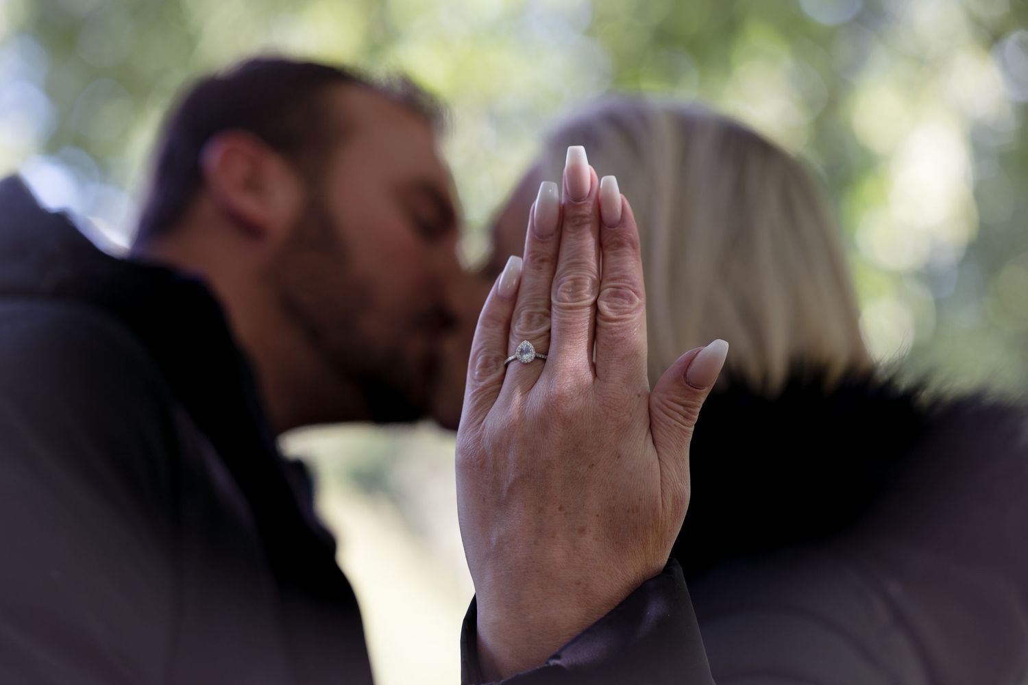 A close-up of a hand displaying an engagement ring while couple kisses in background.