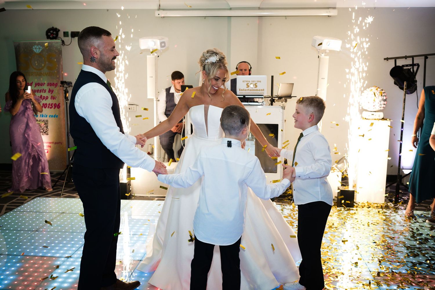 Wedding party guests dancing together in celebration on a brightly lit dance floor at a reception venue.