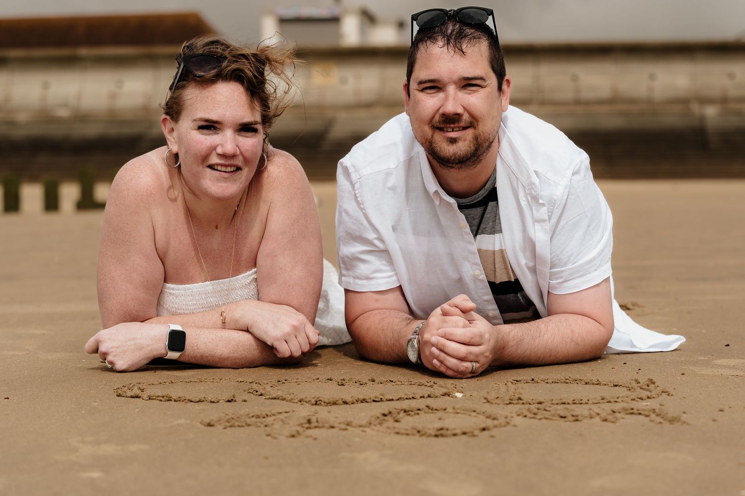 Couple lying on beach sand smiling at camera during a casual photo shoot.
