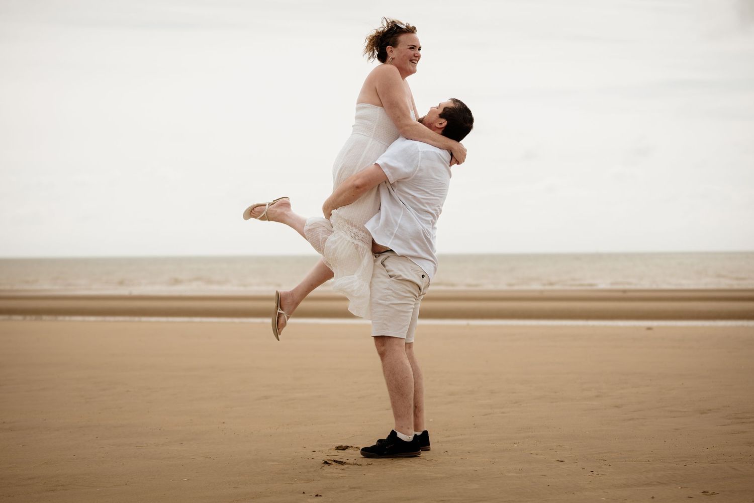 Playful couple embracing on a sandy beach with one person being lifted in the air.