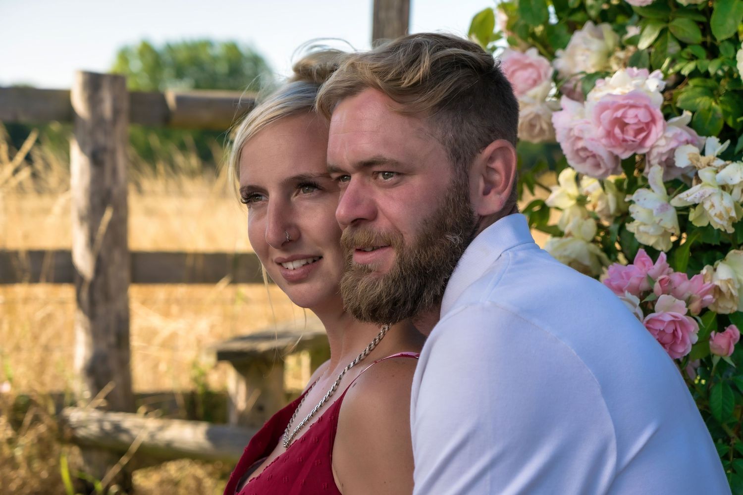 A couple posing together by pink roses and rustic wooden fence during sunset.