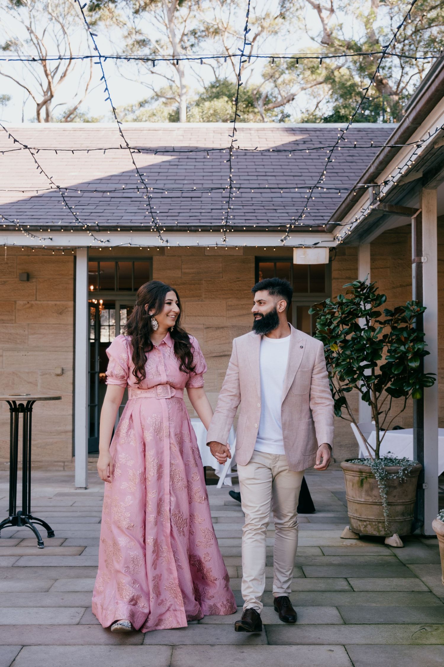 A couple walks hand in hand at a rustic venue, with the person in a pink gown and their partner in a beige blazer.