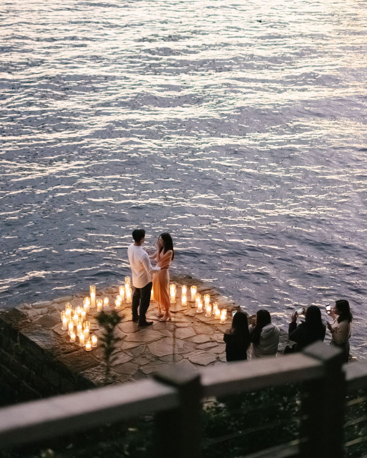 A romantic proposal setup at sunset with candles arranged in a heart shape by the ocean while friends watch from behind.