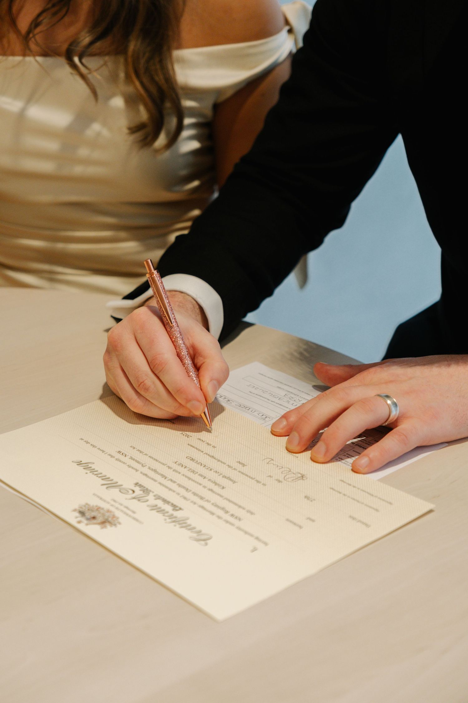 Hands signing a marriage certificate with a gold wedding ring visible on the right hand against a white surface.