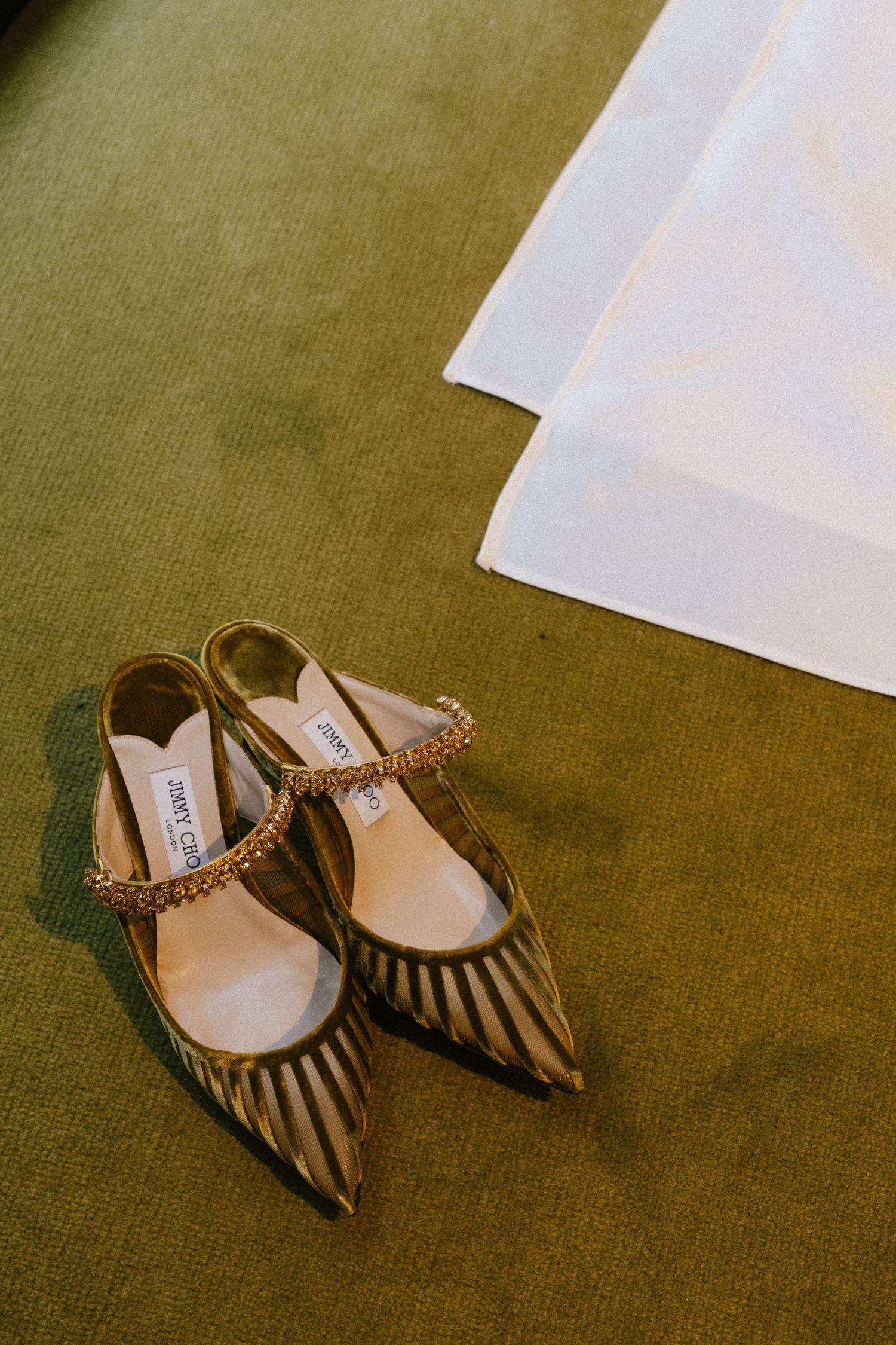 A pair of elegant Jimmy Choo striped slingback heels with studded ankle straps rests on an olive green carpet.