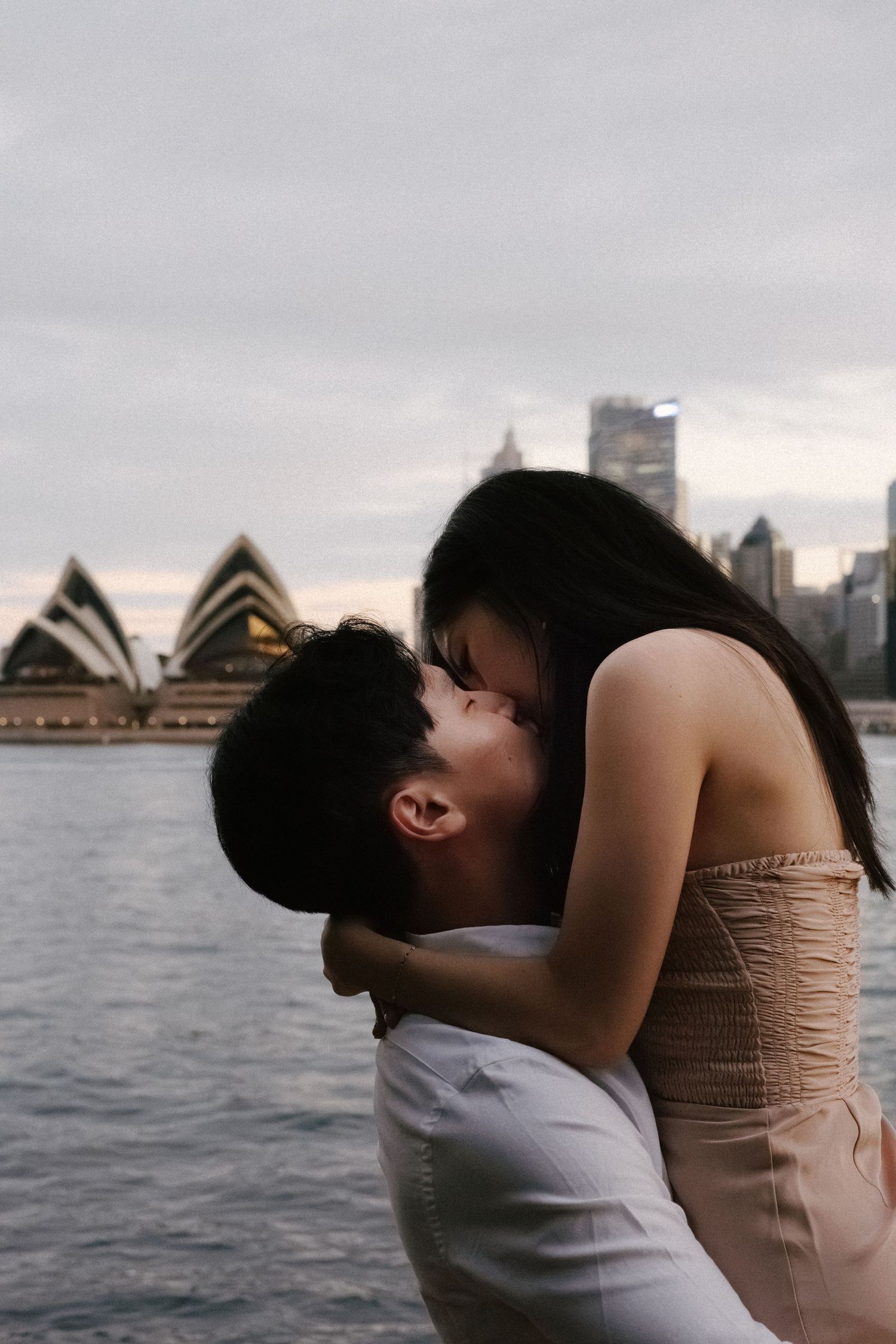 Romantic couple embracing with Sydney Opera House and harbor skyline in background at dusk.