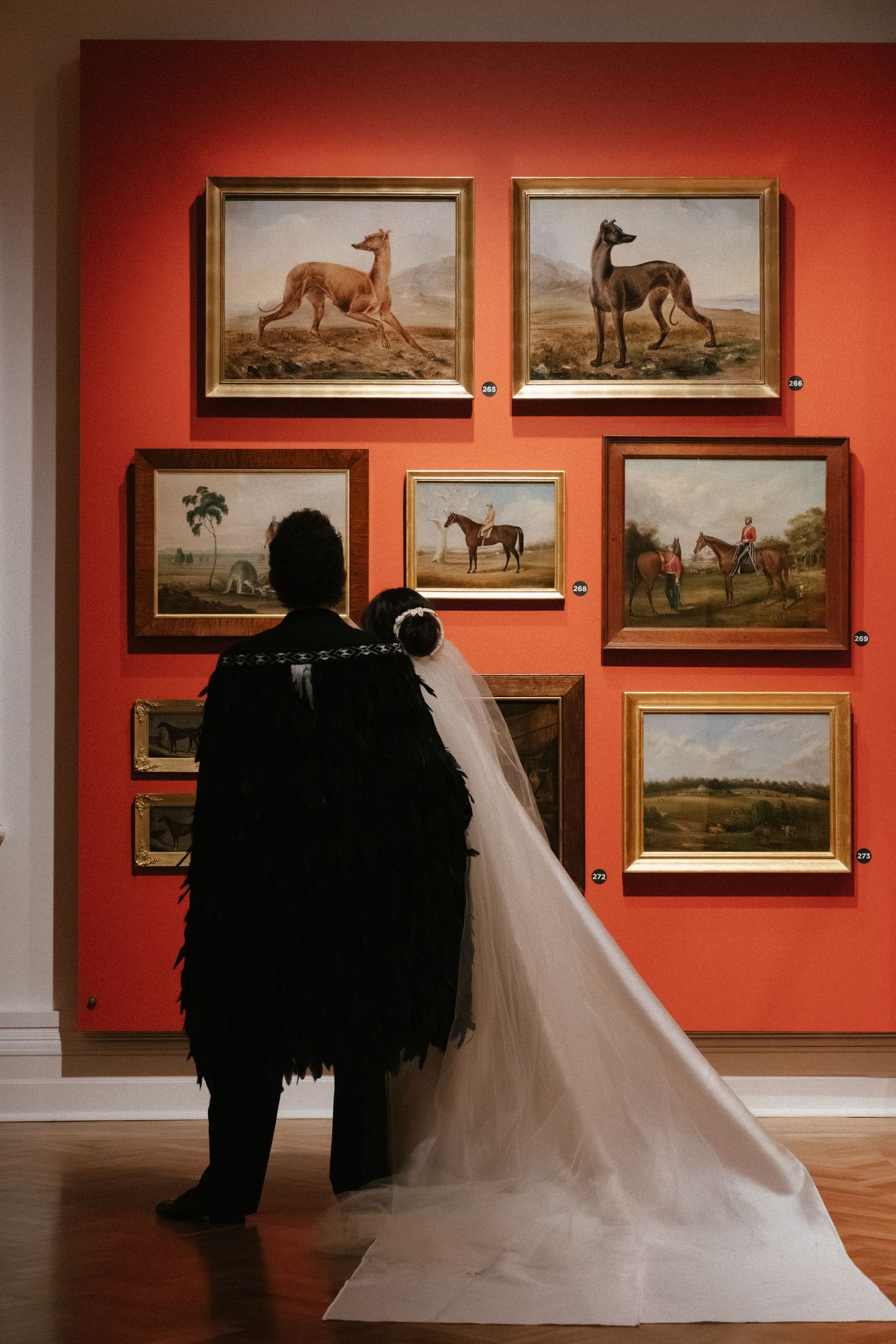 A person wearing a black outfit and white veil views classic paintings of dogs and horses on an orange gallery wall.
