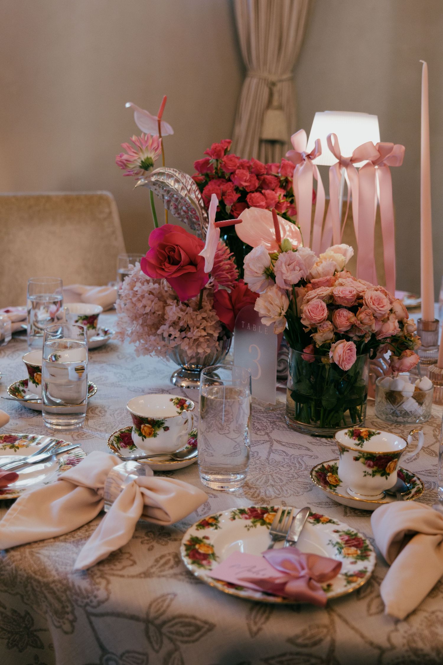 Elegant wedding reception table settings with red and pink flower centerpieces on lace tablecloths under soft lighting.