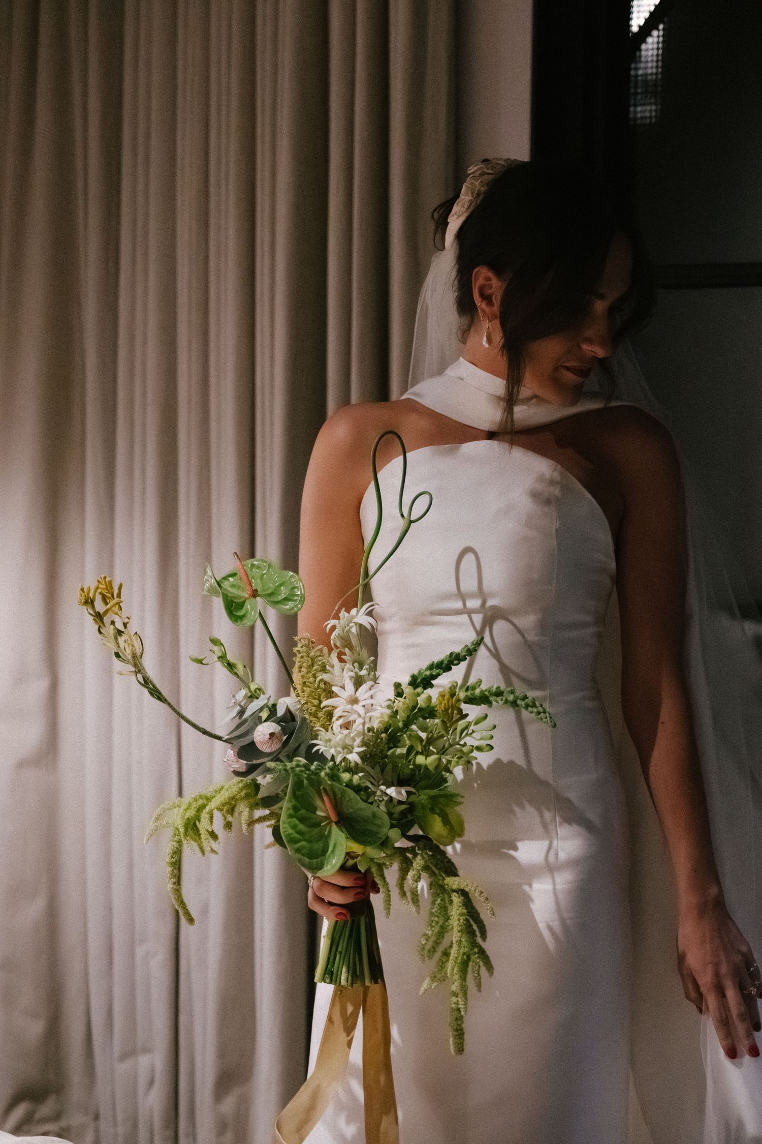 A person in a white wedding dress holds a delicate bouquet of wildflowers against a curtained window background.