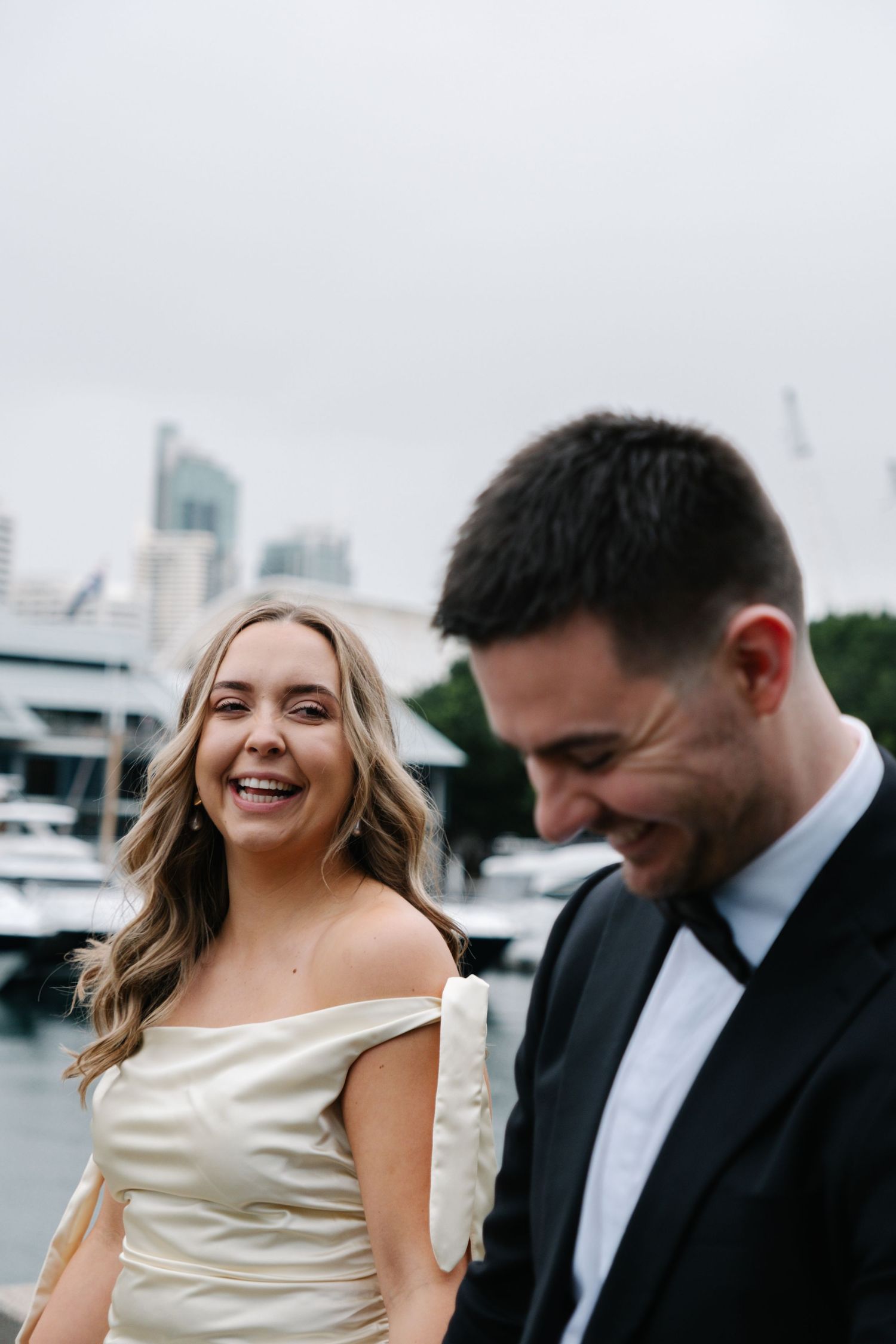 A couple shares a joyful moment together on their wedding day near a marina with city buildings in the background.