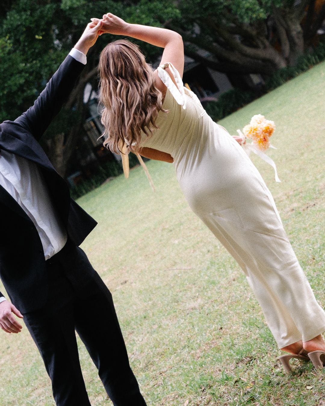 A couple in formal attire dances elegantly on a grassy lawn, with the bride in white and partner in black suit.