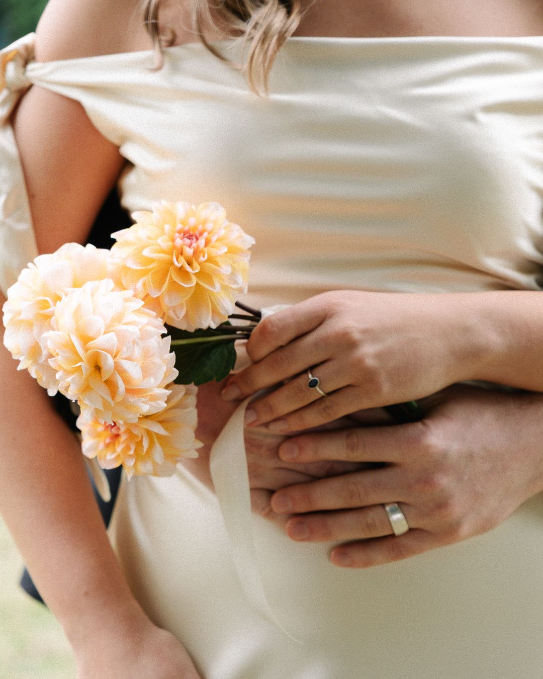 Close-up shot of hands wearing wedding rings while holding delicate peach-colored carnation flowers against white fabric.