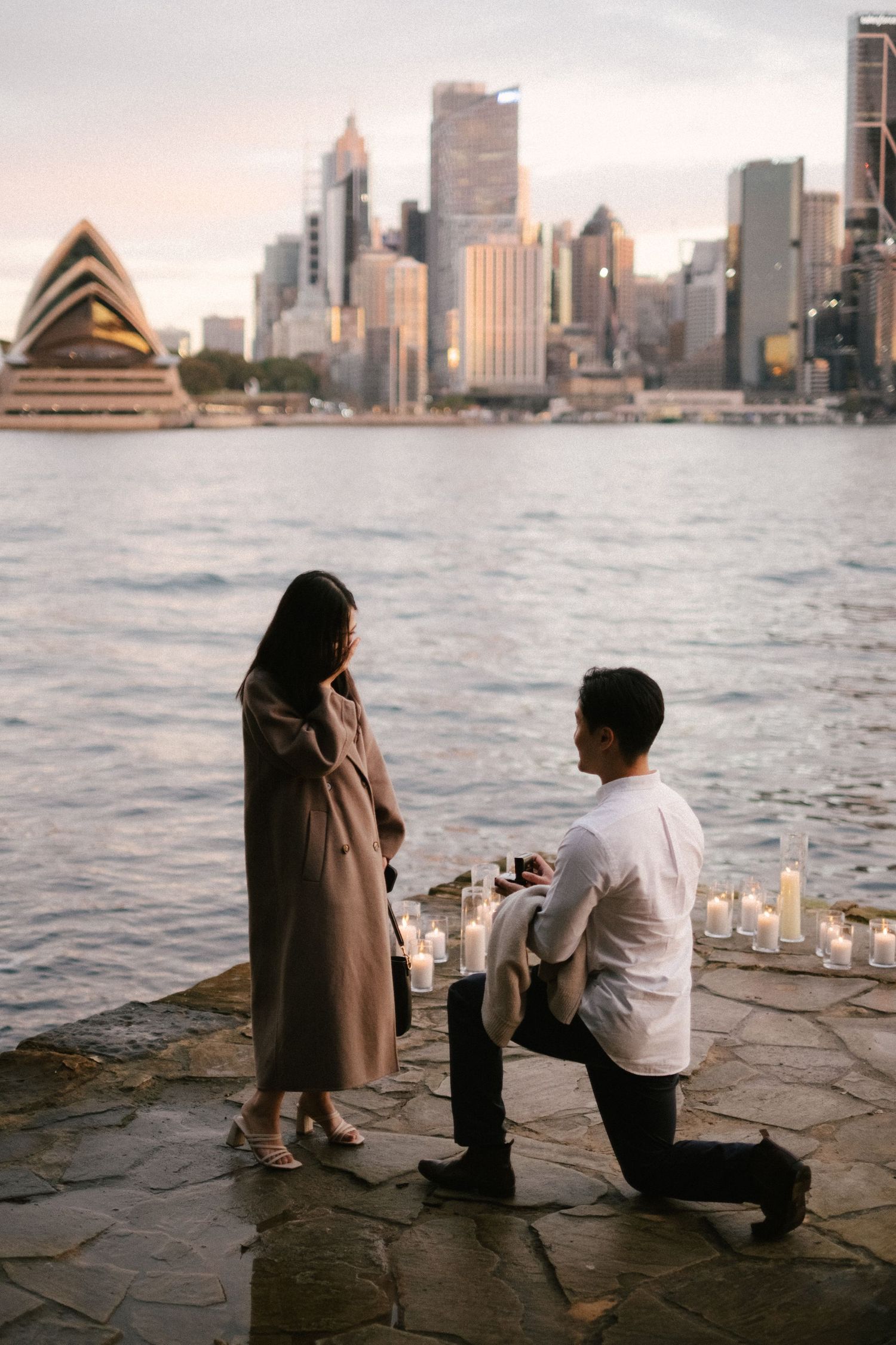A marriage proposal moment unfolds at sunset by the harbor overlooking the Sydney Opera House and city skyline.