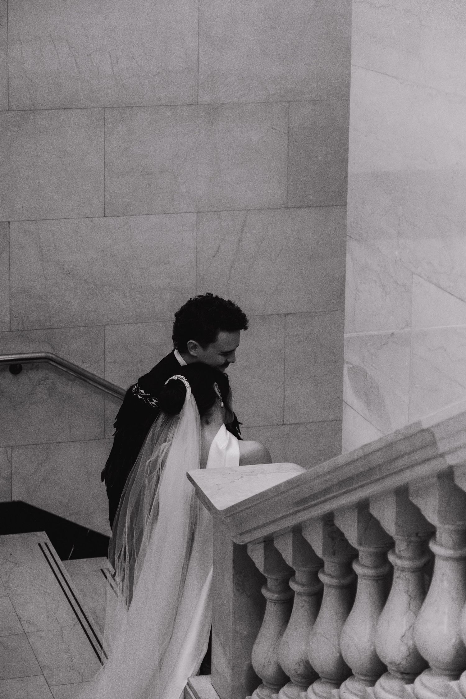 Black and white photo of someone in dark clothing ascending marble stairs with ornate balustrade in elegant interior.
