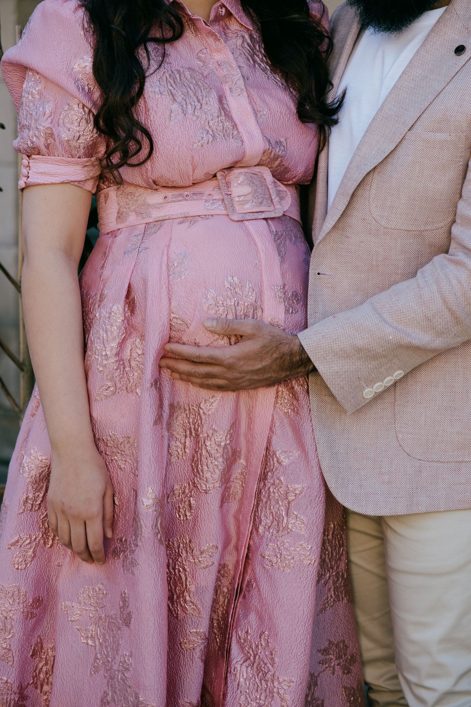 A pregnant person wearing a pink lace dress stands next to someone in a beige suit, with hands gently placed on the belly.