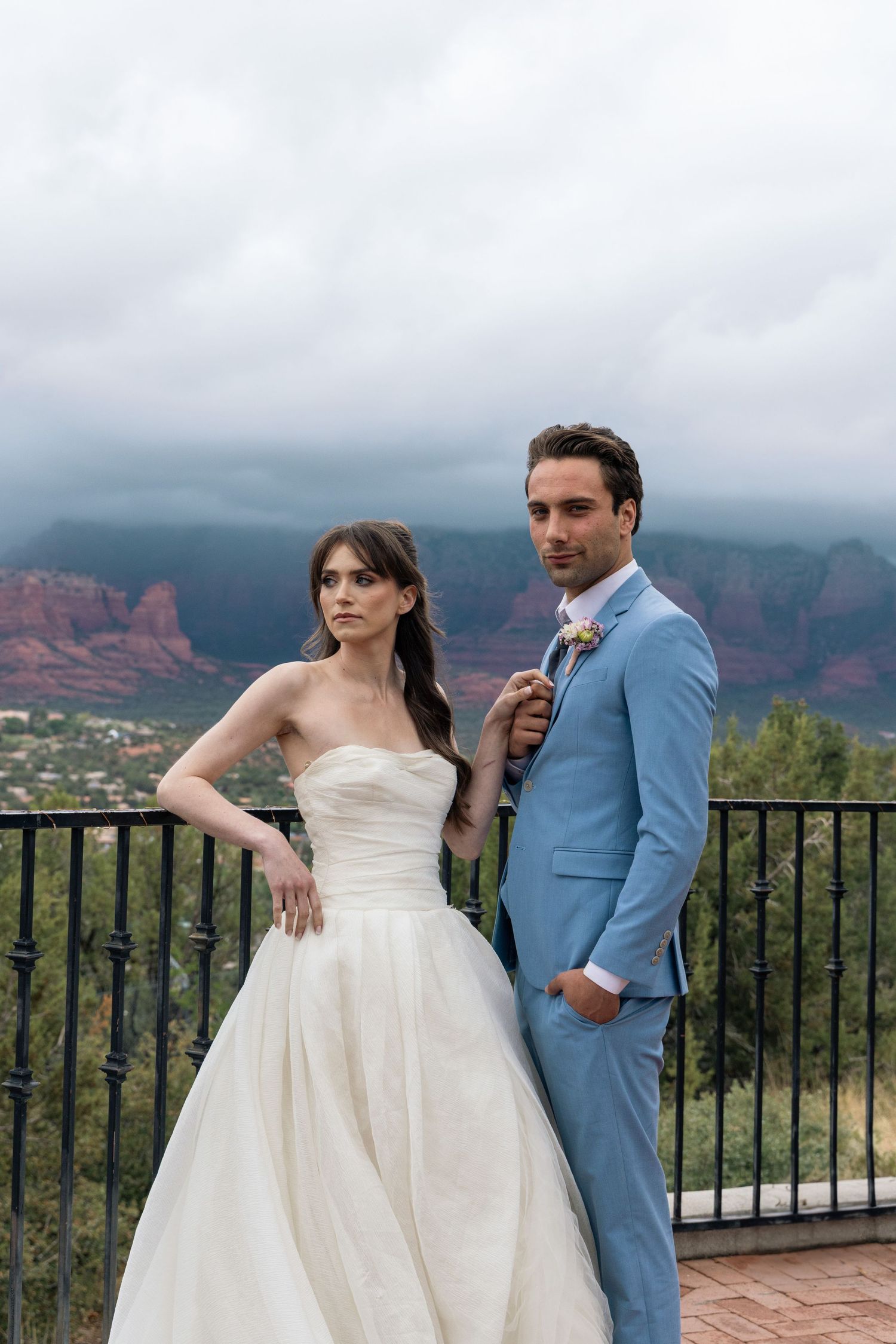 A bride in a white strapless gown and a groom in a light blue suit pose on a balcony overlooking red rock mountains in Sedona.