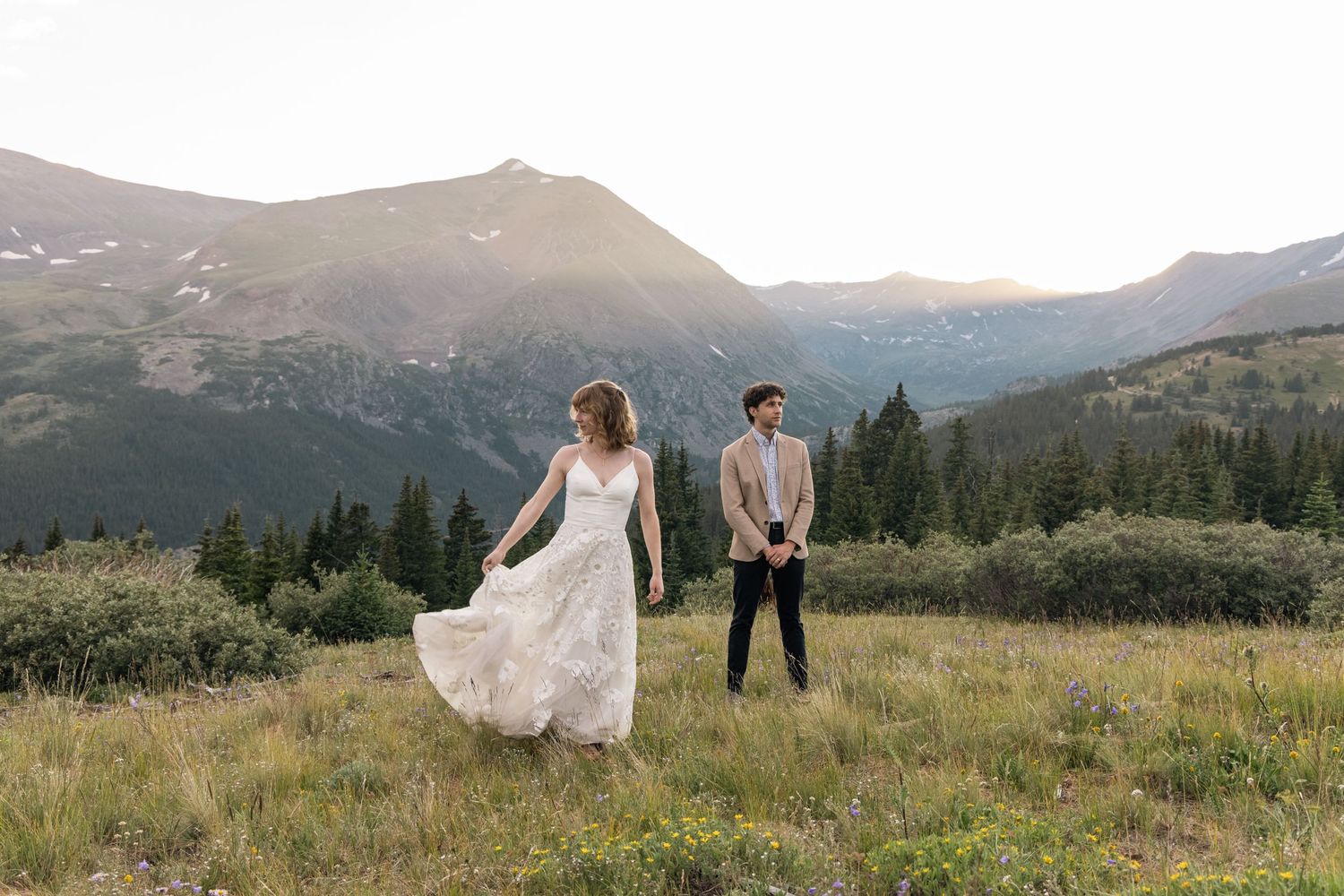 A couple in wedding attire stand in a mountain meadow at sunset with snow-capped peaks in the background.