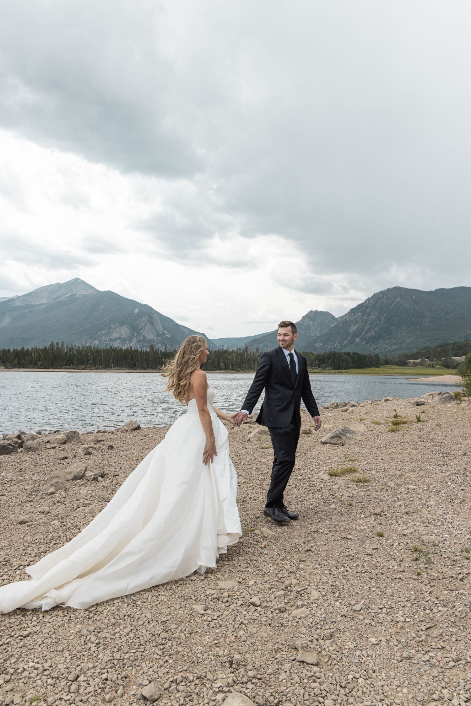 A couple in wedding attire walk hand in hand along a rocky mountain lake shore under dramatic cloudy skies.