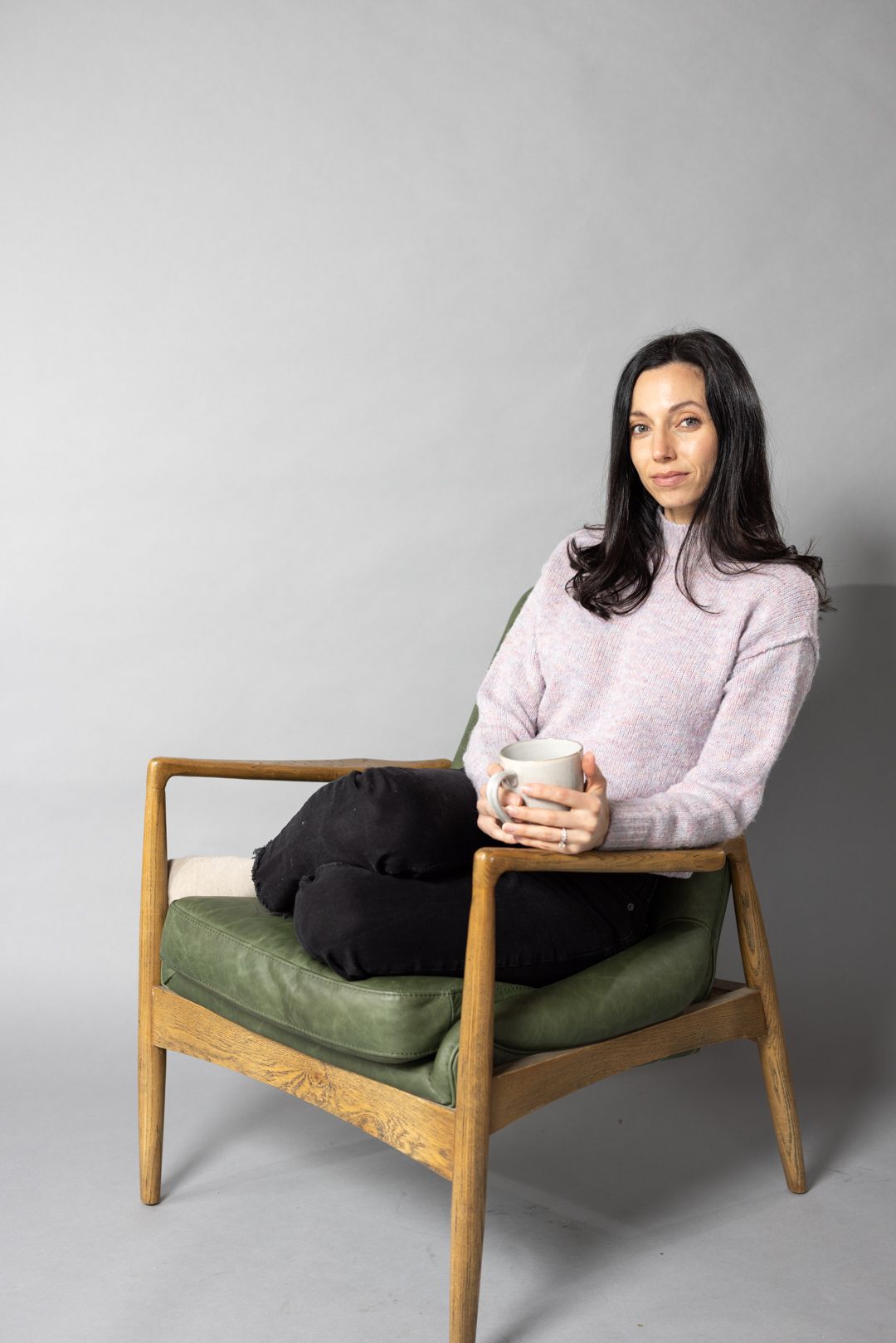 A person in a pink sweater sits on a mid-century modern chair with green upholstery, holding a white mug.