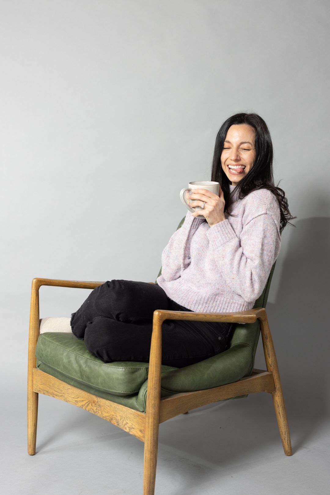 A person lounges in a mid-century armchair with green cushions while holding a warm beverage.