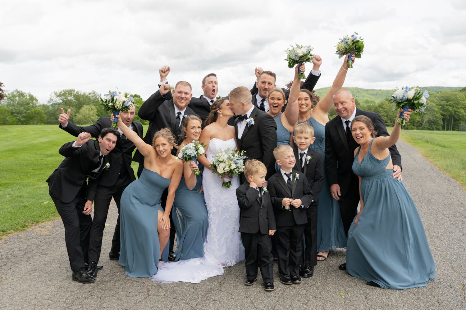 A cheerful wedding party in black suits and dusty blue bridesmaid dresses poses on a country road with arms raised.