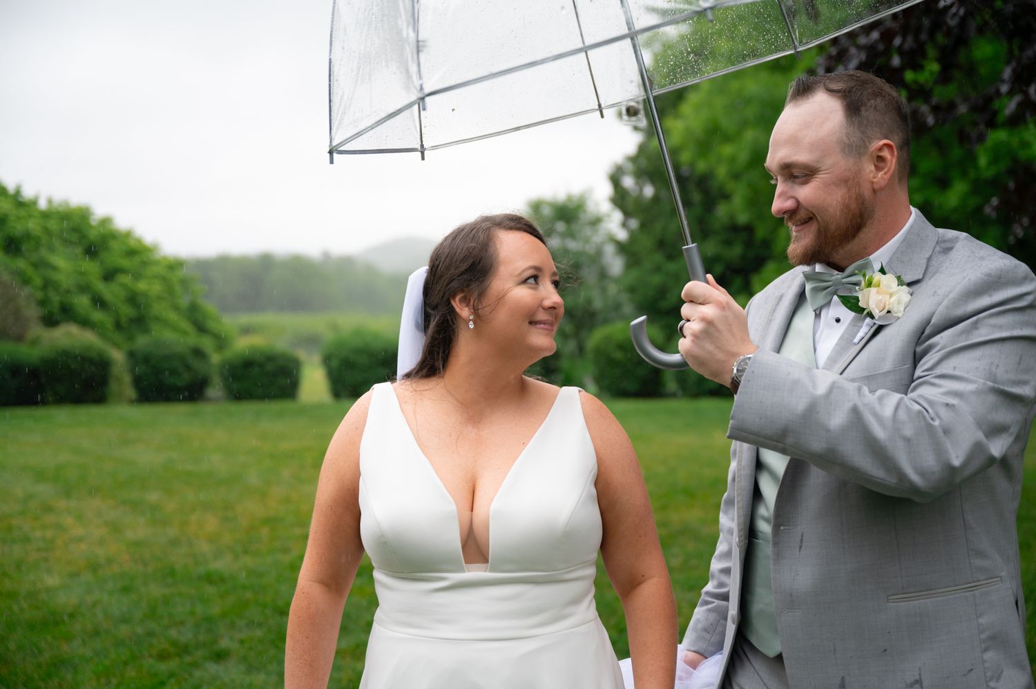 Two people share a joyful moment under a clear umbrella on a lush green lawn during a rainy wedding celebration.