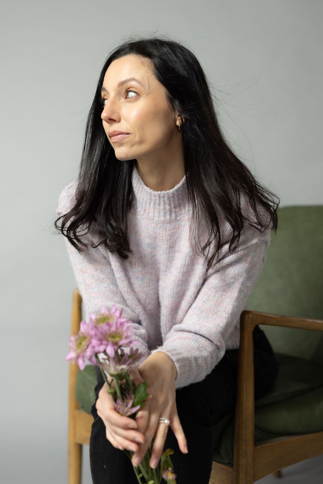 A person in a lilac sweater sits in a wooden chair holding pink flowers against a grey backdrop.